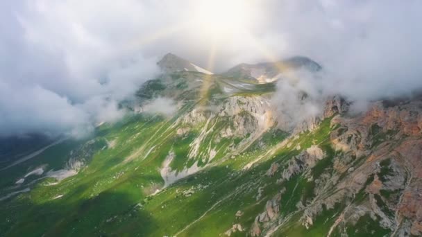 Lotnictwo zbliża się widok na oszałamiające góry Kaukaskie pokryte trawą, śniegiem i kamieniami pod parą ogromnych chmur i jasnych promieni słońca w lecie, Adygea, Rosja. Piękna przyroda z góry. — Wideo stockowe