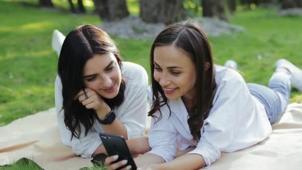 Flickor njuter av fri fritid karaoke i naturen. Para attraktiva positiva flickvänner sjunga sång genom smartphone, ligger på magen på gräs stadspark i vit skjorta och jeans. Närbild — Stockvideo