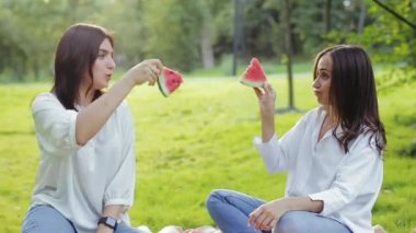 Yakın planda iki güzel kız arkadaşın eğlendiği karpuz dilimleriyle oynadığı, konuştuğu ve şakalaştığı, yazın çimlerin üzerinde oturduğu bir manzara. Hafta sonları arkadaşlarla olumlu aktiviteler.