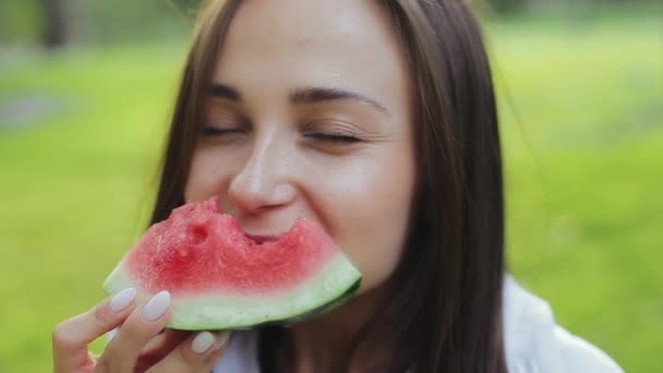Close-up uitzicht van aantrekkelijke schattige brunette jonge vrouw met vriendelijke ogen, snuiven en eten plak watermeloen en glimlachen naar de camera. Mooi en mooi meisje genieten van de smaak van fruit buiten in park. — Stockvideo