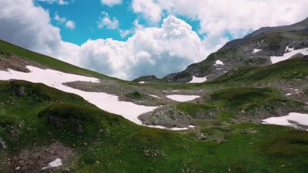 Pandangan atas lereng gunung Kaukasia yang menakjubkan di bawah awan melonjak dari atas di langit dalam cuaca cerah, sebagian tertutup oleh lubang dengan salju di pagi musim panas. Terbang di atas alam Adygea, Rusia. — Stok Video