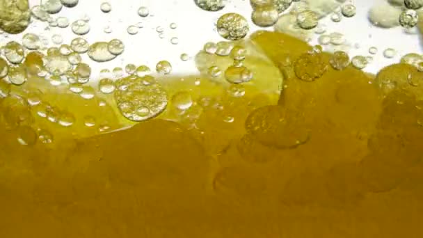 Close-up van gouden vloeistof in een glas. Vloeibare gele golflijn. Midden uitzicht op grote en kleine luchtbellen op een witte achtergrond. Goudkleurige vloeistof in een container. Zonnebloemolie gemengd met water — Stockvideo