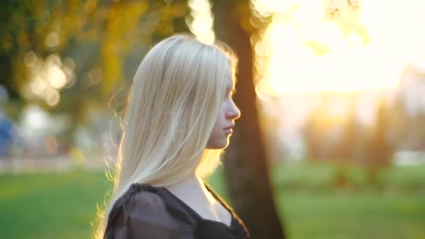 Attraktiv ung blond flicka i snygg svart klänning promenader i varm våren park mot en bakgrund av gräs och träd och tittar på kameran med en lockande utseende, filmisk närbild i profil. — Stockvideo