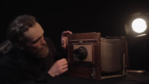 Бородатый человек снимает крышку старой деревянной ретро-аккордеонной камеры, регулирует объектив под светом лампы в темно-изолированной комнате. Проверка редкого устаревшего оборудования, концепция восстановления антикварной камеры . — стоковое видео