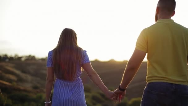 Zadní pohled na milující mladý běloch pár krásné dívky a vousatý chlap drží ruce v poli na trávě a těší nádherný večerní západ slunce ve zpomaleném filmu. Romantické datum a volný čas v přírodě. — Stock video