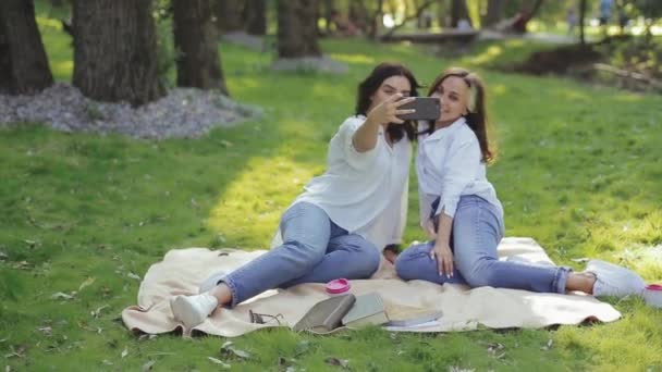 Twee positieve en aantrekkelijke meisjes nemen verschillende selfies in prachtige poses, zittend op bedsprei over gras in zonnig stadspark tussen de bomen. Actieve vrije tijd buiten in de frisse lucht van mooie vriendinnen. — Stockvideo