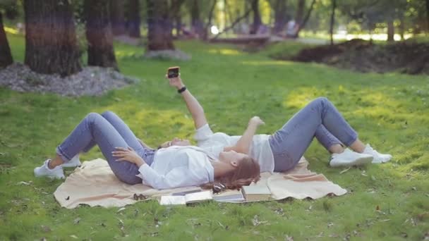 Paire de jolies copines souriantes prennent selfies couchés dans des directions opposées côte à côte sur l'herbe sur couvre-lit dans un parc confortable parmi les arbres. Deux belles jeunes femmes positives passent des loisirs actifs. — Video