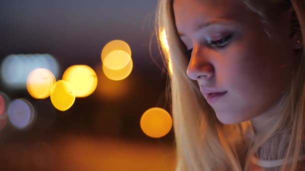 Carino sorridente bionda felice in abito bianco chatta con gli amici su smartphone sullo sfondo di luci sfocate di auto in serata, vista da vicino. Passeggiata notturna nelle luci della città. Concetto di tecnologia urbana — Video Stock