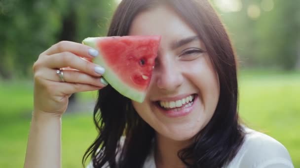 Lachend aantrekkelijk brunette meisje van Europese verschijning kijkt door plak van watermeloen en eet met plezier lachen op camera en eten in zomerpark. Begrip actieve levensstijl en vrijetijdsbesteding. — Stockvideo
