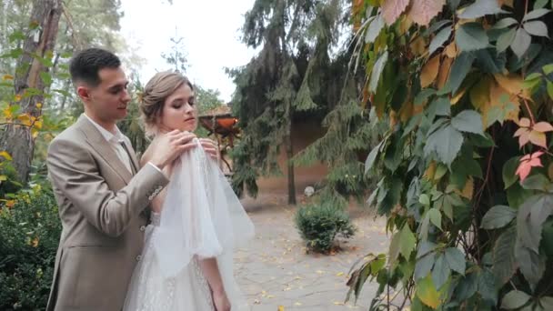 Vista de cerca de la feliz pareja de boda europea en la naturaleza. Novio en traje abraza a la novia y endereza su vestido de novia en su hombro contra el fondo de abetos y una casa en el bosque . — Vídeo de stock