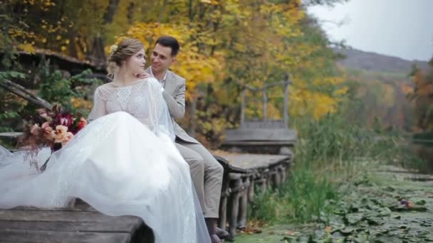 Vista media de la feliz pareja de boda besándose y abrazándose junto al río mientras se sienta en un puente de madera cerca del bosque de otoño. Luna de miel romántica para los encantadores recién casados. Linda novia y novio disfrutar de vacaciones. — Vídeo de stock