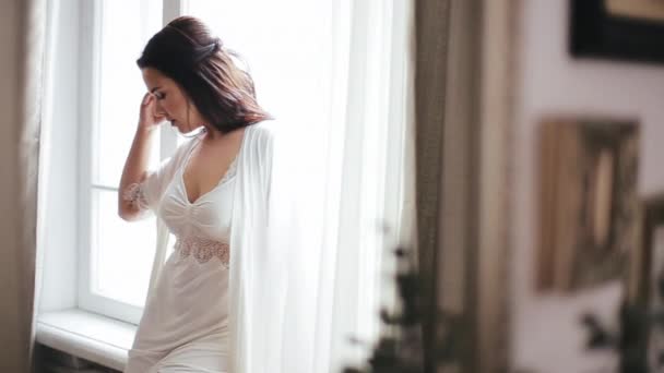 Novia joven caucásica con cabello castaño en un lujoso peignoir blanco se encuentra con una nueva mañana del día en el alféizar de la ventana del dormitorio, alisando su cabello y peignoir. Sexy mujer europea por la ventana . — Vídeos de Stock