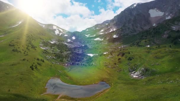 Вид з повітря на гору, вкриту щільною зеленою травою, сніг з невеликим озером з каменів і дерев під сонячними променями і райдужною веселкою влітку. Ландшафт природи краси — стокове відео