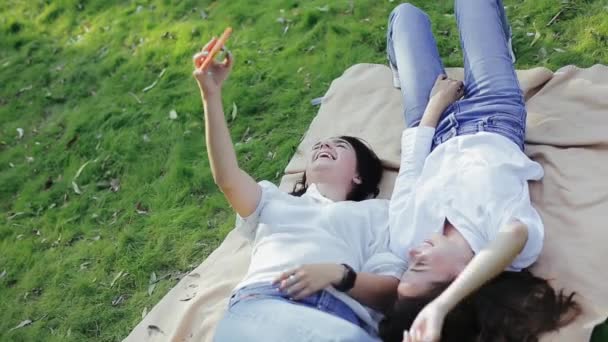 Duas garotas ridentes alegres tomam selfie no telefone, deitado na grama no parque na manhã de primavera ensolarada. Conceito de tempo de lazer na natureza, estilo de vida ativo. Meninas bonitas gostam ativamente de descansar fora — Vídeo de Stock