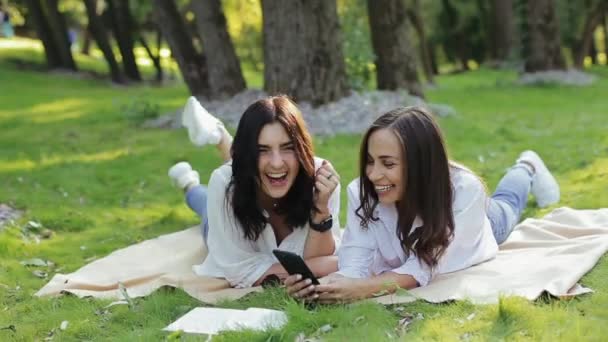 Δύο γελαστά όμορφα κορίτσια να κείτονται στο γρασίδι του πάρκου, συζητώντας για selfie στο smartphone. Μερικά όμορφα κορίτσια διασκεδάζουν ενεργά. Έννοια ενεργό απολαμβάνοντας τον τρόπο ζωής, ελεύθερο χρόνο στη φύση — Αρχείο Βίντεο