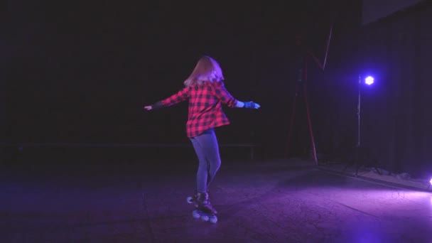 スローモーションでスポットライトの光の下で放棄された建物の暗いシーンで前輪に交差脚を持つ1つの場所でローラーに回転する美しい若いブロンドの女の子。アクティブなライフスタイルの概念 — ストック動画