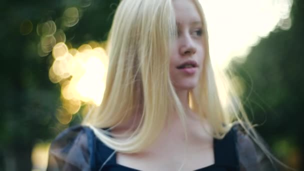 Charmant jong Europees blond met lang haar en een zwarte jurk kijkt weg en dan in de camera met een verleidelijke mooie blik tegen de achtergrond van de felle zon en bomen in het stadspark. — Stockvideo