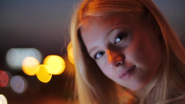 Cara jovem sorridente atraente de loira europeia sorrindo bonito para a câmera contra o fundo de lanternas luminosas brilhantes da cidade da noite. Fotografia de uma linda garota bonita com cabelo comprido . — Vídeo de Stock