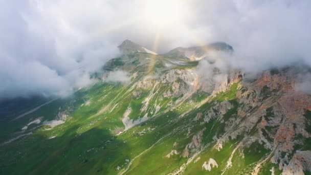 Vue aérienne approchant des superbes montagnes du Caucase couvertes d'herbe, de neige et de pierres sous la vapeur d'énormes nuages et des rayons de soleil brillants en été, Adygea, Russie. Belle nature d'en haut. — Video