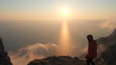 Sportif erkek turist yüksek bir dağın kenarına doğru koşar ve deniz üzerinde gün batımının büyüleyici güzellikteki manzarasına ve bulutların dumanına hayran kalır. Aktif yaşam ve dağ ve kayalarda yürüyüş.