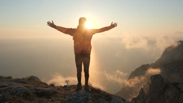 Člověk stojí na okraji vysoké hory nad mořem, roztahuje paže na boky a vlny a zakrývá si tělo zářícím sluncem, zblízka. Vousatý turista stojí při západu slunce u útesu. — Stock video