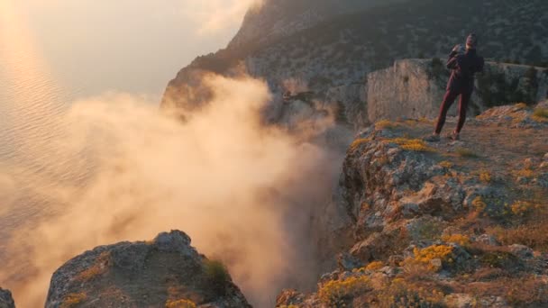 Une jeune femme sportive filme diverses vapeurs et fumées provenant de nuages sous une haute montagne, debout au bord d'une falaise sur fond de magnifique coucher de soleil sur la mer dans la soirée. — Video