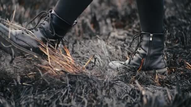 Vue rapprochée des jambes féminines dans les leggings dans des chaussures élégantes en cuir à semelle haute qui se tiennent sur le sol de cendres et de coups de pied dans l'herbe noire brûlée après le feu de champ par temps ensoleillé de printemps au ralenti. — Video