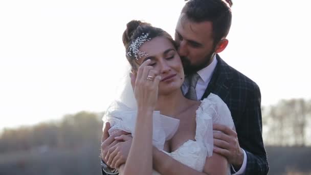 Vue rapprochée de beaux jeunes mariés passant du temps ensemble dans la nature par une chaude journée ensoleillée d'automne. Joyeux marié câlins et bisous de derrière sa belle mariée dans une robe sur fond de champ. — Video