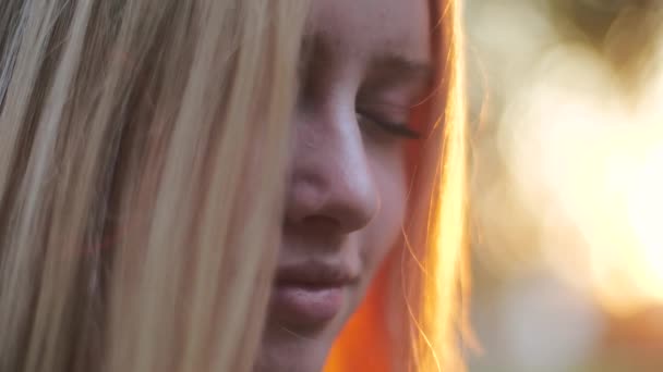 Retrato de cerca de una adolescente europea pacificada disfrutando del aire fresco y el sol dorado de color suave al atardecer. El viento sopla sobre el cabello rubio. Belleza natural, emociones verdaderas. Ternura, concepto de feminidad — Vídeos de Stock