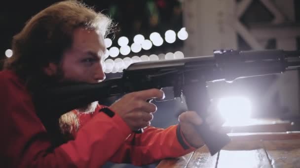 Un homme millénariste caucasien aux cheveux blonds et barbus tire depuis une mitrailleuse dans un champ de tir tard dans la nuit, un personnage masculin tient une arme dans ses mains, vise, se penche sur une boîte, lumières en arrière-plan . — Video