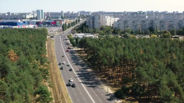 Górny widok ruchu drogowego na drodze asfaltowej przechodzącej między lasem sosnowym na tle budynków mieszkalnych i nieba w słoneczny dzień w Charkowie, Ukraina. Zdjęcie lotnicze podróżujących samochodów i pojazdów. — Wideo stockowe