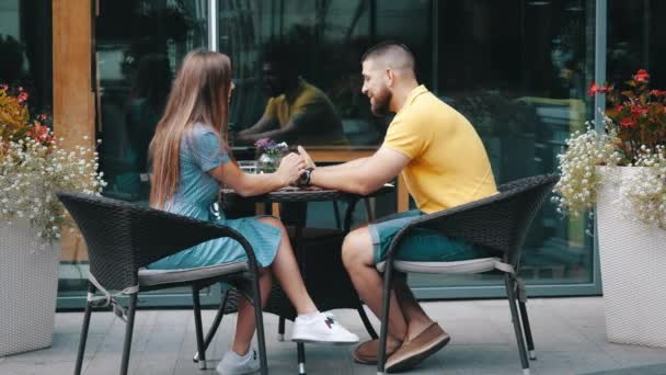 Joven linda pareja en el amor chico y chica en ropa urbana sentado en la mesa de la cafetería afuera y hablando, tomados de la mano en cámara lenta. Cita romántica en restaurante al aire libre. Desayuno por la mañana en el establecimiento — Vídeos de Stock