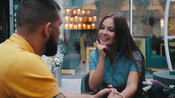 Vista de cerca de la hermosa niña sonriente sentada en la mesa y hablando al aire libre de la cafetería de moda con novio barbudo, de la mano, fondo de velas detrás de la ventana. Cita romántica hombre y mujer — Vídeos de Stock