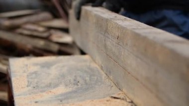 Kimliği belirsiz bir marangozun çalışma eldivenleriyle yakın plan görüntüsü. Kalın bir tahtayı tekrar birleştiriyor ve onu öğütücüyle nasıl keseceğini merak ediyor. Adam dairesel makineyle kesmek için tahta parçayı inceliyor..