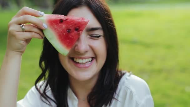 Mooie en vrolijke jonge brunette vrouw maakt verschillende grappige grimaces met plak watermeloen, eet het in het park op wazige achtergrond van gras. Actieve recreatie van grappig meisje met fruit in het weekend. — Stockvideo