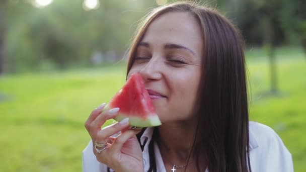 Gelukkig mooi brunette meisje in wit shirt geniet van snuiven plak watermeloen met plezier eten en lachen, zitten in gezellige zomerpark. Weekend actief vrijetijdsconcept met fruit buiten in de frisse lucht. — Stockvideo