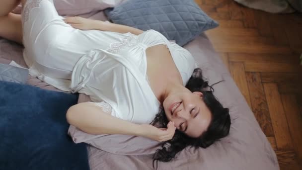 Belle jeune fille européenne la mariée en sous-vêtements blancs et peignoir se trouve sur le lit dans la pose sexuelle et profite de la matinée, souriant de plaisir avec les yeux fermés. Séance photo en studio de mariée mignonne — Video