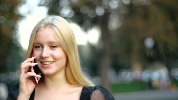 Uzun sarı saçlı neşeli beyaz kız öğrenci dışarıda telefonda konuşurken eğleniyor, mutlu bir şekilde gülümsüyor. Akrabalar ve arkadaşlarla iletişim, aşk ve romantizm, boş zaman ve rahatlama. — Stok video