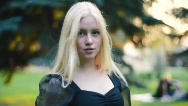 A young girl in a black dress in slow motion confidently walks through the park and looks at the camera, smiling attractively. The concept of femininity and health, leisure and outdoor activities. — Stock Video