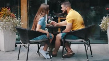 Genç ve mutlu çiftin öpüşmelerinin orta görüntüsü. Yaz sabahları, modern açık hava kafesinde el ele tutuşuyorlar. Yavaş çekimde. Sakallı adam masadaki kızla iletişim kuruyor. Erkek ve kadın randevuda..