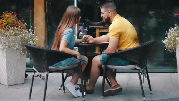 In der Mitte ein junges glückliches verliebtes Paar, das sich küsst und Händchen haltend an einem Sommertag in Zeitlupe in einem modernen Café im Freien steht. Der bärtige Kerl kommuniziert mit dem Mädchen am Tisch. Mann und Frau auf Date. — Stockvideo
