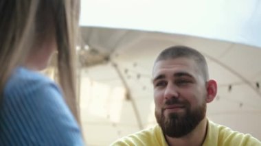 Yüzü sakallı, sarı polo giyen, dışarıda, kafede oturan ve kızların sohbetini dinlemekten zevk alan kısa saçlı adamın yakın görüntüsü. Aşık genç çiftin romantik eğlencesi..