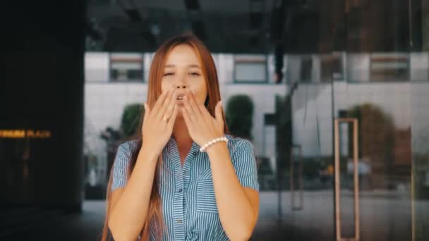 Vista frontal de la encantadora linda chica de pelo largo sonriente en vestido a rayas enviando besos de aire a la cámara contra la pared reflectante de vidrio del restaurante. Positivo feliz joven mujer transmite besos de amor . — Vídeos de Stock