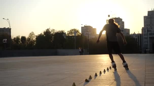 Mladý dlouhosrstý muž bruslař tancuje mezi kužely na večerním náměstí při západu slunce. Freestyle slalom Roller bruslař ve zpomaleném filmu. Stylový městský chlap tráví volný čas ve sportu — Stock video