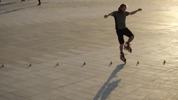 Man rullskridskoåkare med långt hår dans mellan kottar gör rotationer på kvällen stad vid solnedgången. Freestyle slalom Boy rullskridskor i slow motion. Snygg urban kille tillbringar sin lediga tid inom sport — Stockvideo