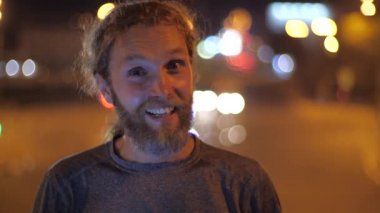 Gülümseyen yakışıklı, beyaz sakallı, uzun saçlı genç adam doğrudan kameraya bakar ve hareket halindeki bir şehrin arka planında baş parmağını gösterir..