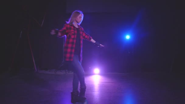 Charmant blond meisje in beschermende handschoenen op rolschaatsen begint professioneel spinnen op voorwielen met benen gekruist in donkere scène onder licht van schijnwerpers. Atletische en actieve levensstijl. — Stockvideo