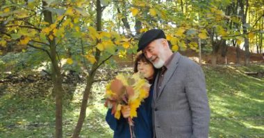 Parkta birbirine sarılıp gülümseyen, yakın çekim yapan olgun bir çift. Bir buket yaprakla büyüleyici emeklilerin karısı güneşli bir havada parktaki ağaçların arasında sonbahar tatilinin tadını çıkarıyor..