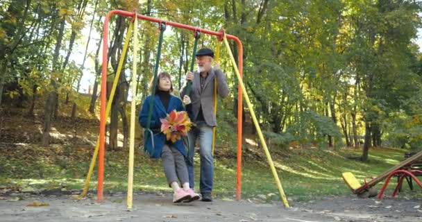 Baard man schudt een schommelstoel met zijn vrouw die een boeket herfstbladeren vasthoudt. Levendig actief bejaardenpaar dat zich vermaakt in een stadspark. Swnior familie van gepensioneerden veel plezier in de buitenlucht. — Stockvideo