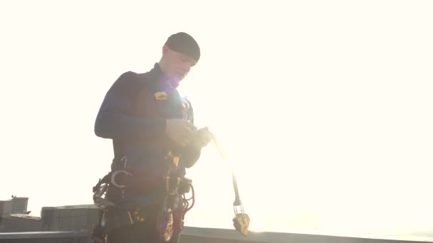 Vista de cerca de un escalador masculino profesional en un uniforme en la azotea que asegura cierres y ganchos con correas en el cuerpo para cable y seguro. Una profesión peligrosa para los hombres valientes y extremos . — Vídeo de stock